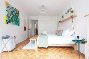 A bedroom with a bed, a hanging light fixture, a wooden bench, a small table, and a wall-mounted shelf with plants.