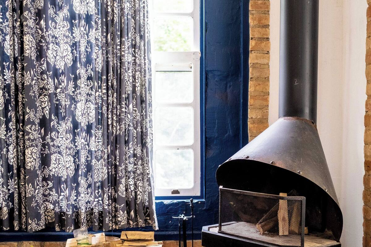 Une pièce avec une cheminée, une fenêtre avec des rideaux et une table ornée d’un vase de fleurs.