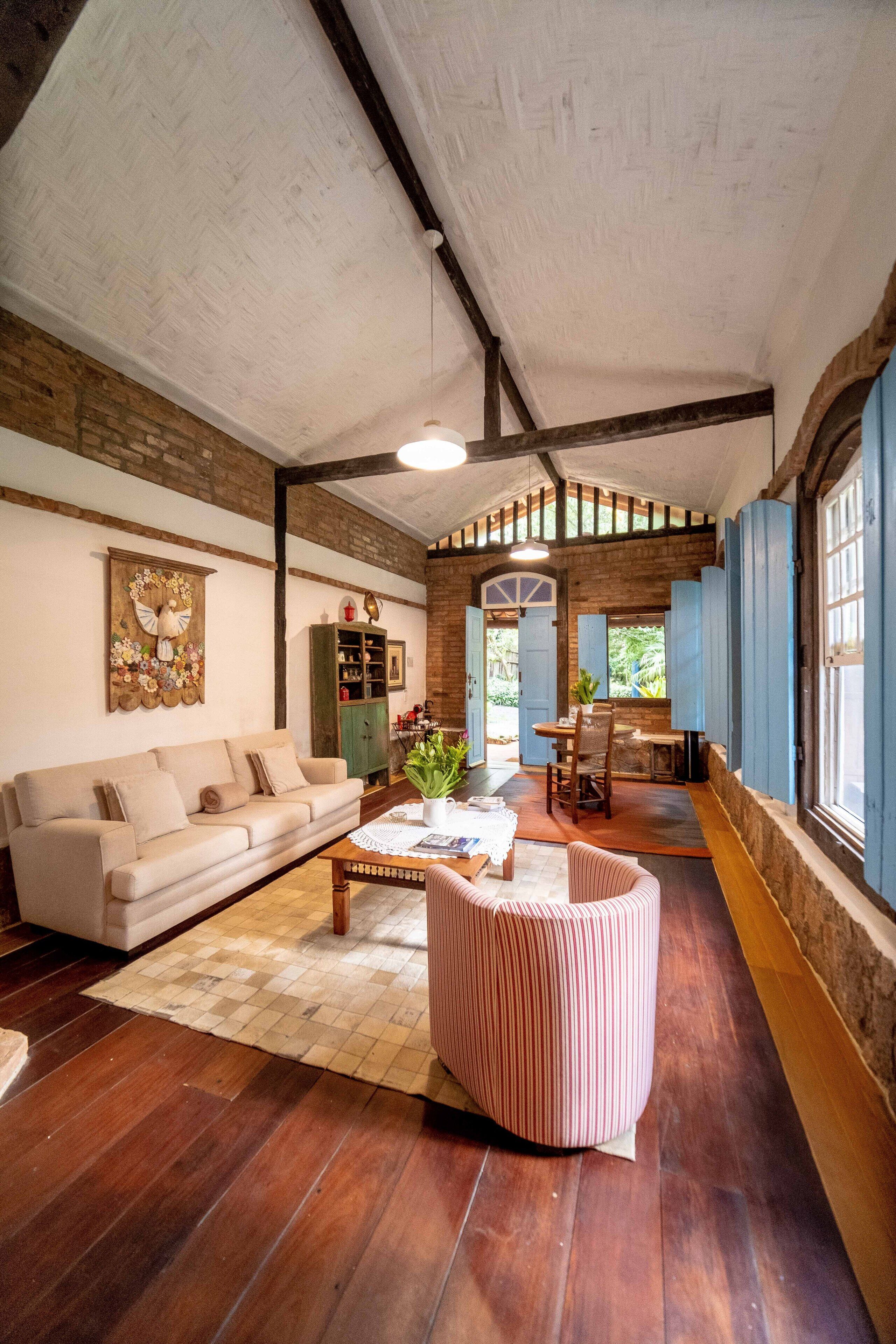 A spacious living room with a high ceiling, wooden beams, a beige sofa, a coffee table, and a striped ottoman.