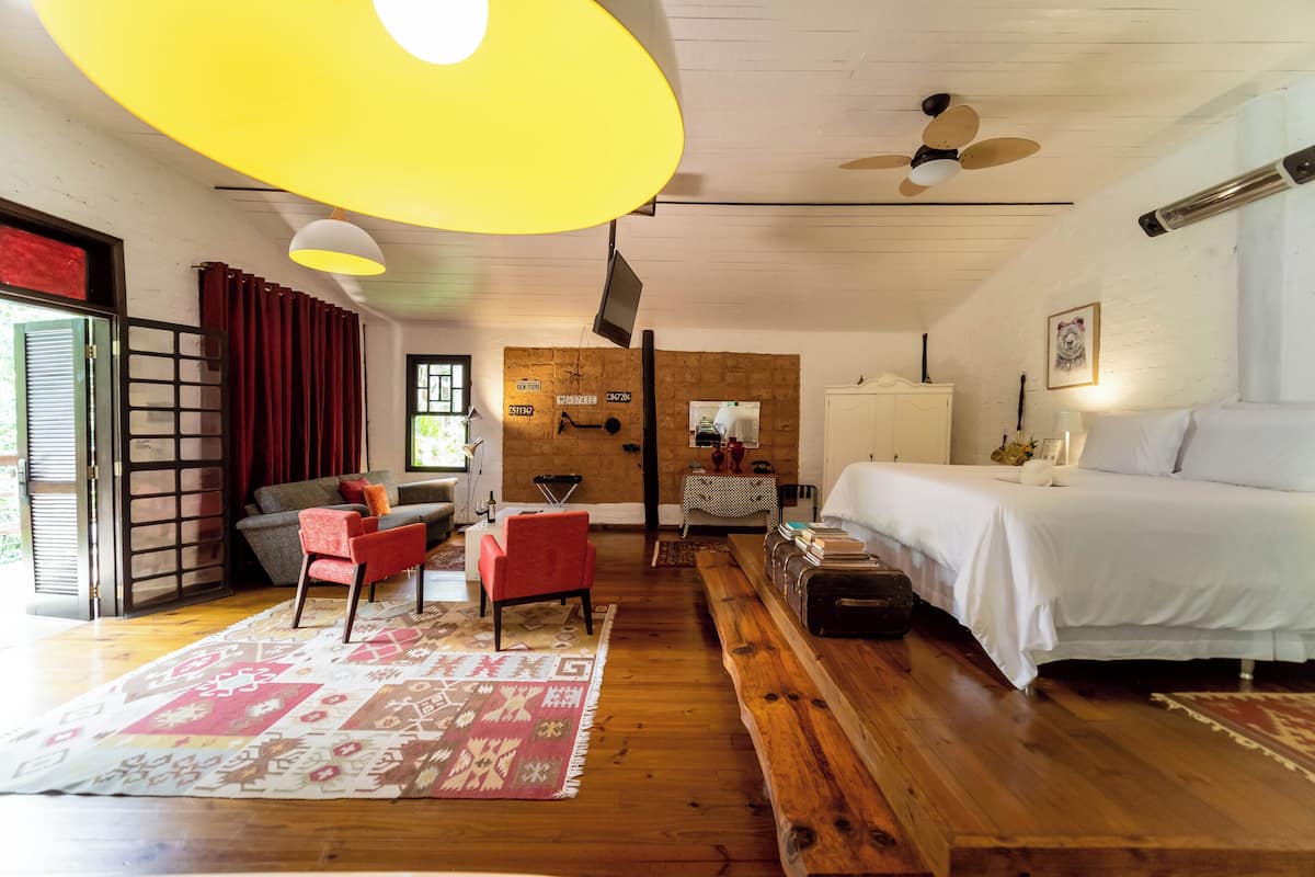 Une pièce comprenant un lit, un canapé, un fauteuil rouge, un banc en bois, un tapis à motifs et un ventilateur de plafond.