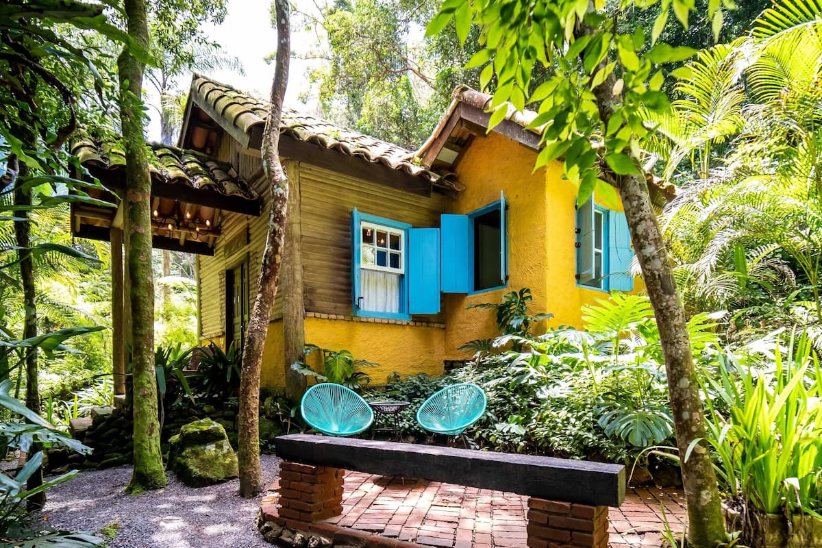 Une maison à l’extérieur jaune et aux volets bleus, entourée d’une végétation luxuriante et bordée d’un chemin de gravier.