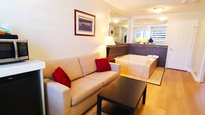 A compact living space with a kitchenette, a sofa with red cushions, a small table, a bathtub, and a mirror reflecting the room. - Riviera Inn and Suites 1000 Islands (Gananoque)