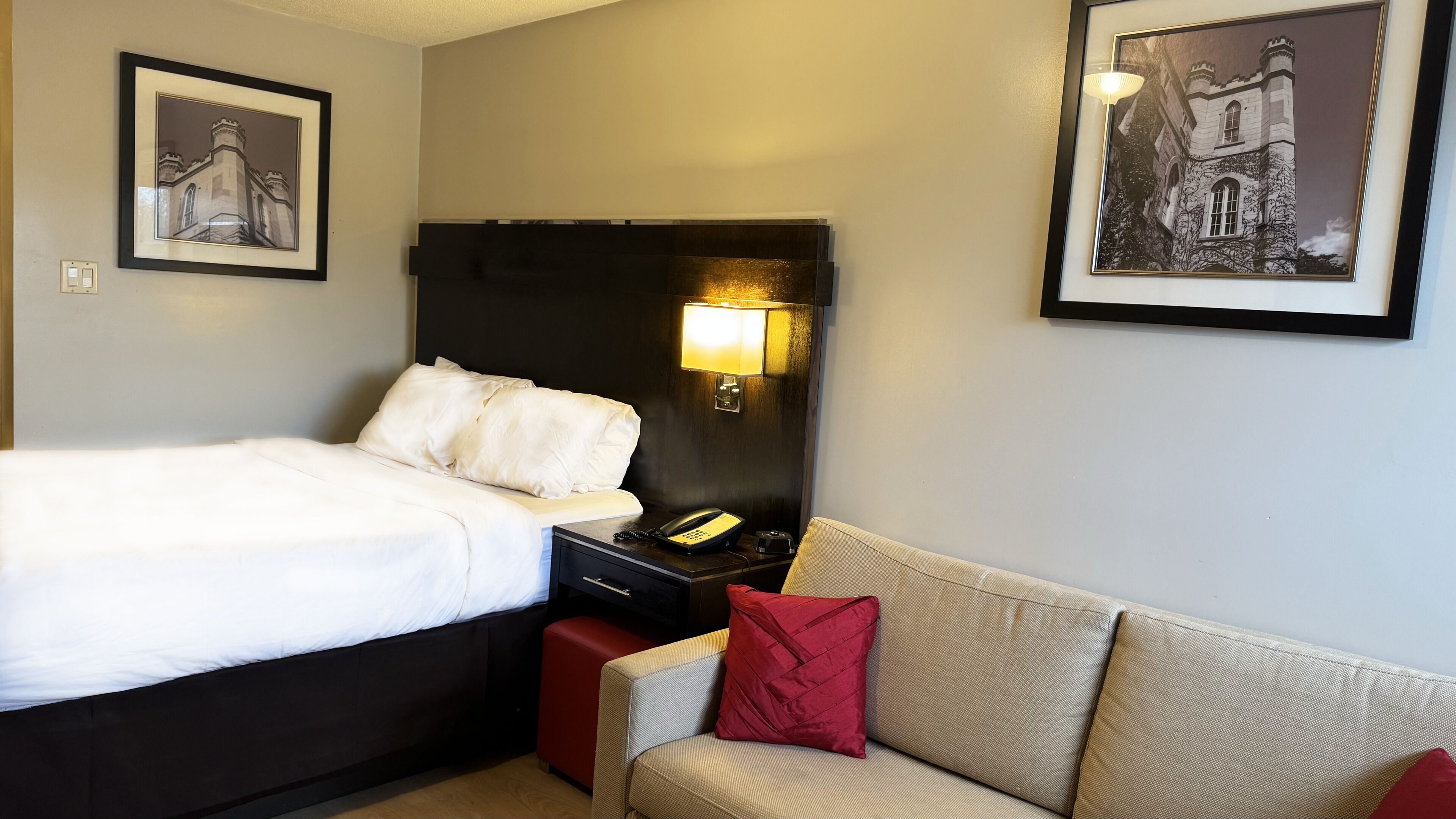 A hotel room with a bed, a nightstand with a lamp, a telephone, and a couch with a red pillow.