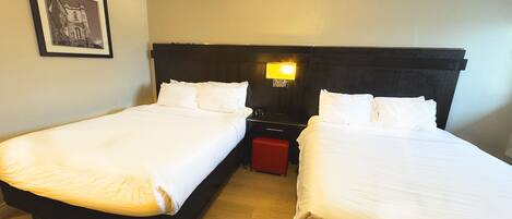 Habitación de hotel con dos camas individuales, una mesita de noche con lámpara, un cuadro en la pared y un banco rojo.