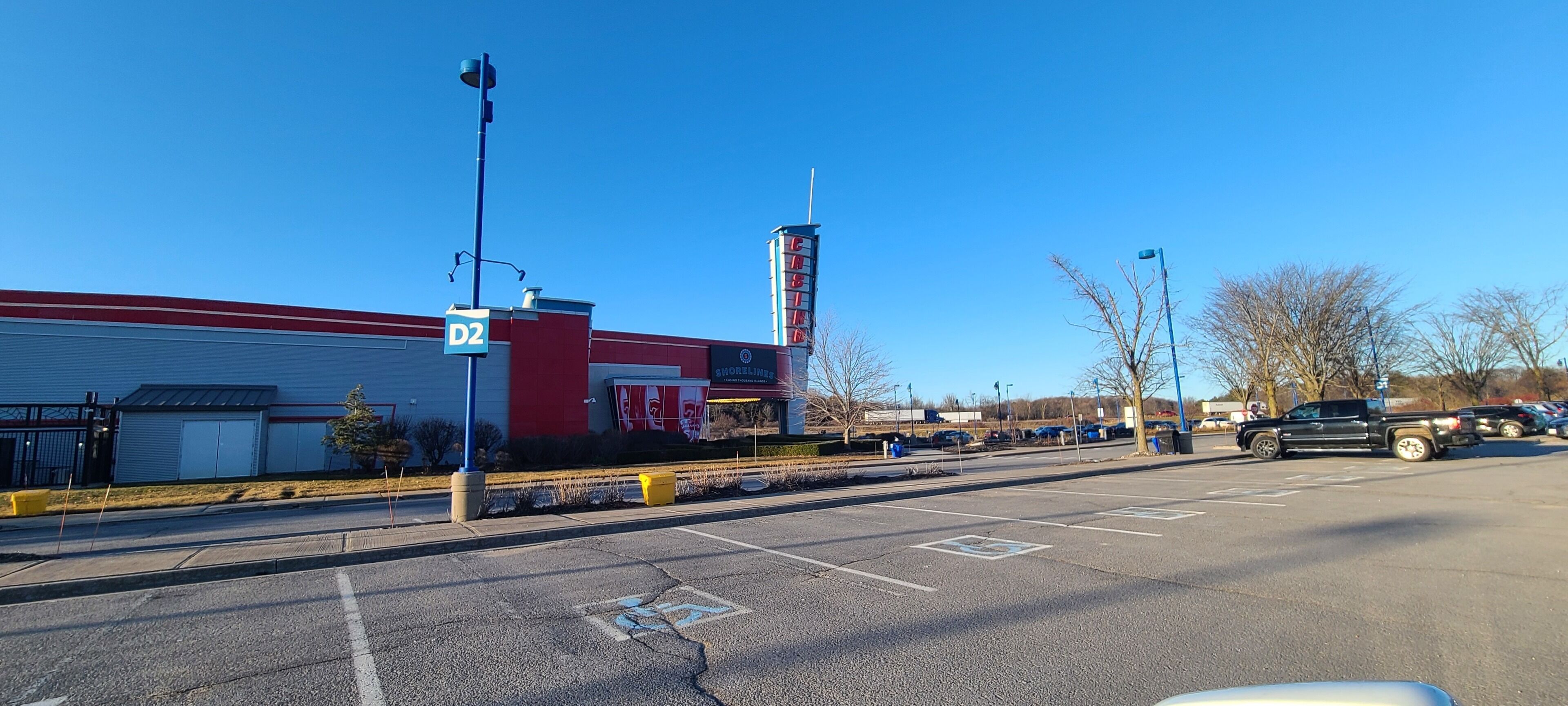 A parking lot with a D2 sign, a building with a red and white facade, and a tall structure in the background.