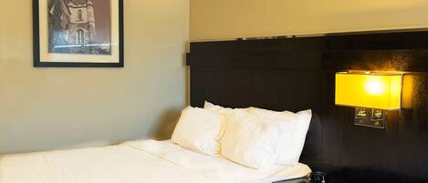 Een hotelkamer met een bed, een nachtkastje, een digitale klok, een lijst met foto aan de muur en een lamp bevestigd aan de muur.