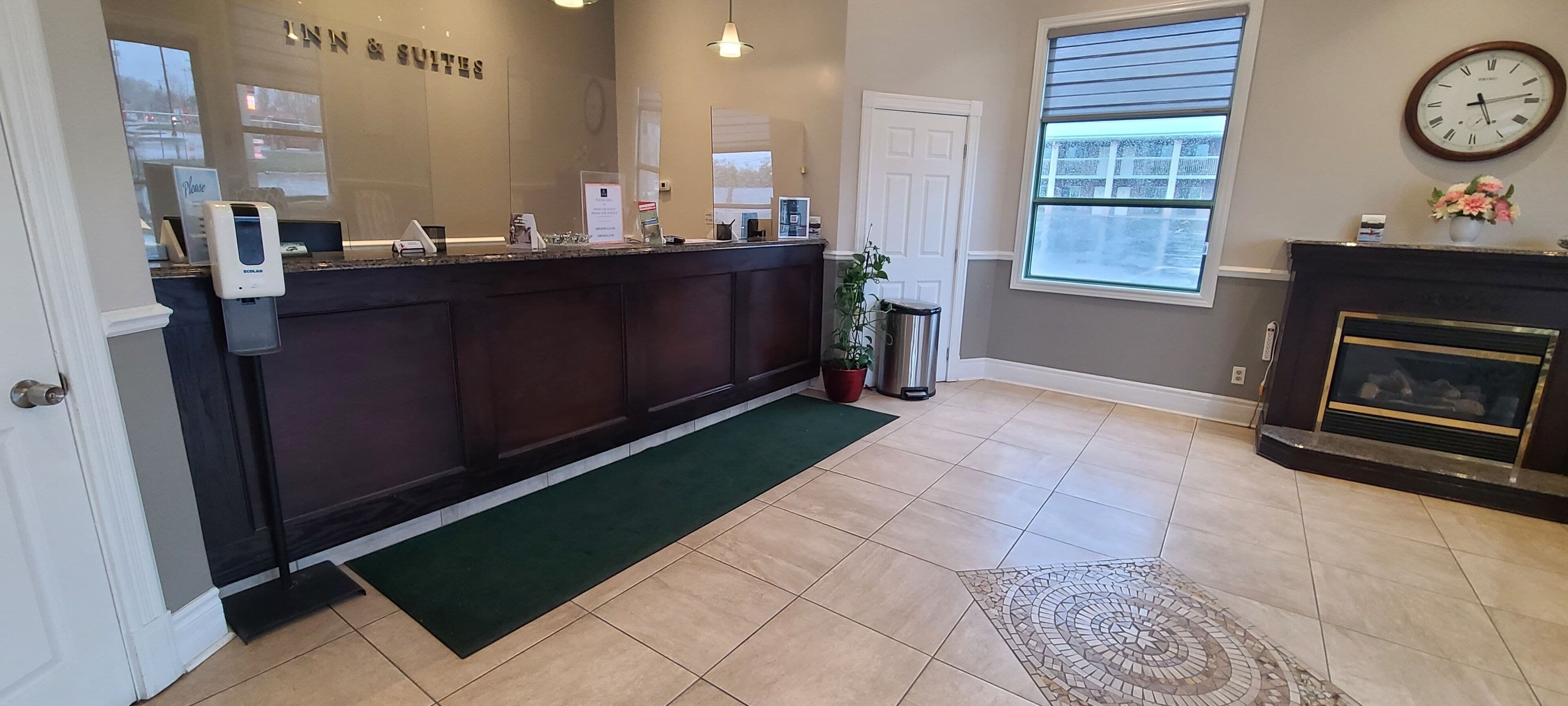 A hotel reception area with a reception desk, a clock, a fireplace, and a window with blinds.