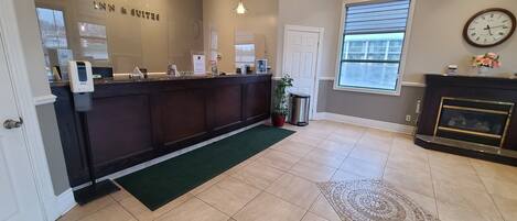 A hotel reception area with a reception desk, a clock, a fireplace, and a window with blinds.