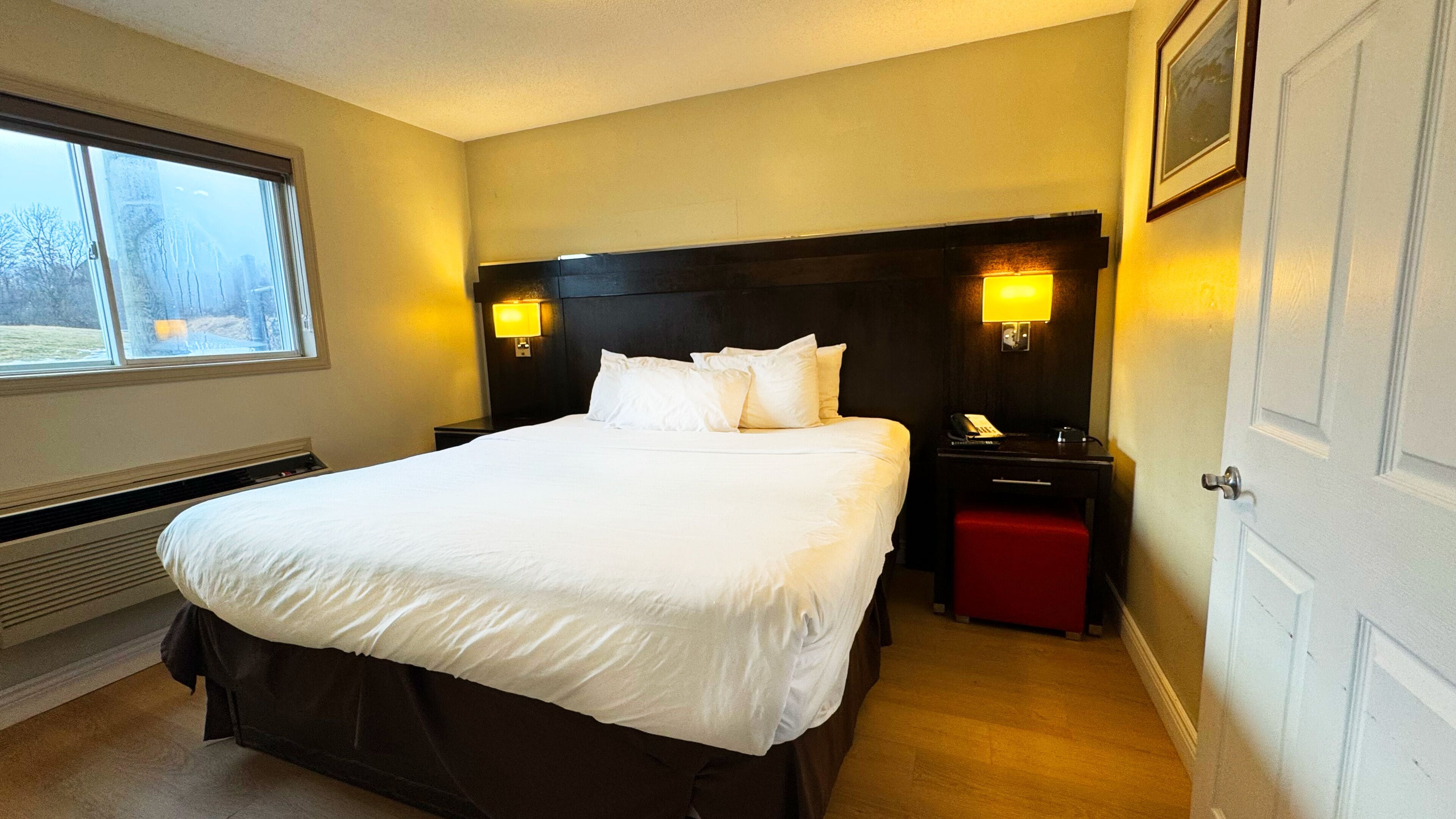 A hotel room with a large bed, two bedside lamps, a nightstand, a red ottoman, a heater, and a window with a view of trees.