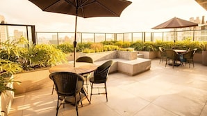 A rooftop terrace with outdoor seating, including a table and chairs, surrounded by planters and a large umbrella.