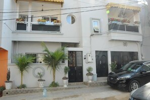 Un immeuble résidentiel de deux étages doté de balcons, avec une voiture noire garée devant et des plantes en pot disposées le long de l’entrée.