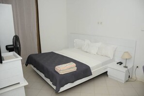 Un lit blanc avec une couverture et des oreillers foncés, une table de chevet avec une lampe et une petite commode blanche.