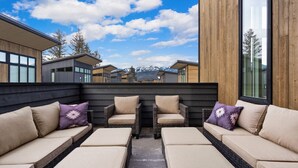 A modern outdoor seating area with cushioned sofas and chairs, a wooden deck, and a view of mountains in the background. - New! Meticulously Designed 3BR, 3.5B with Pool, Hot Tub, Fire Pit Access! (Whitefish)