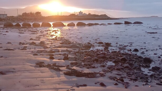A beach with rocks and sand, a sunset in the background, and a distant shoreline.
