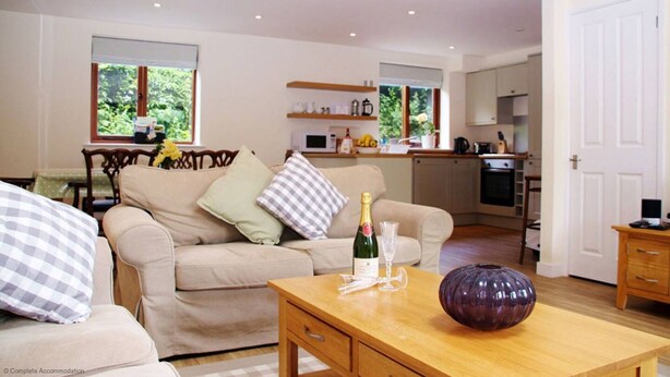 A living room with a sofa, a wooden coffee table with a bottle and glass, and a kitchen in the background. - Middledown -  a cottage that sleeps 7 guests  in 3 bedrooms (Newton Valence)