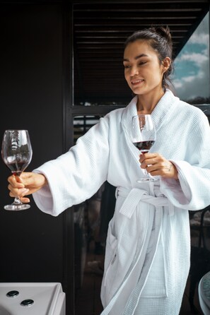 A person in a white bathrobe holding a wine glass, standing in a room with a dark wall and a window with a view. - Gaia glamping  (San Rafael)