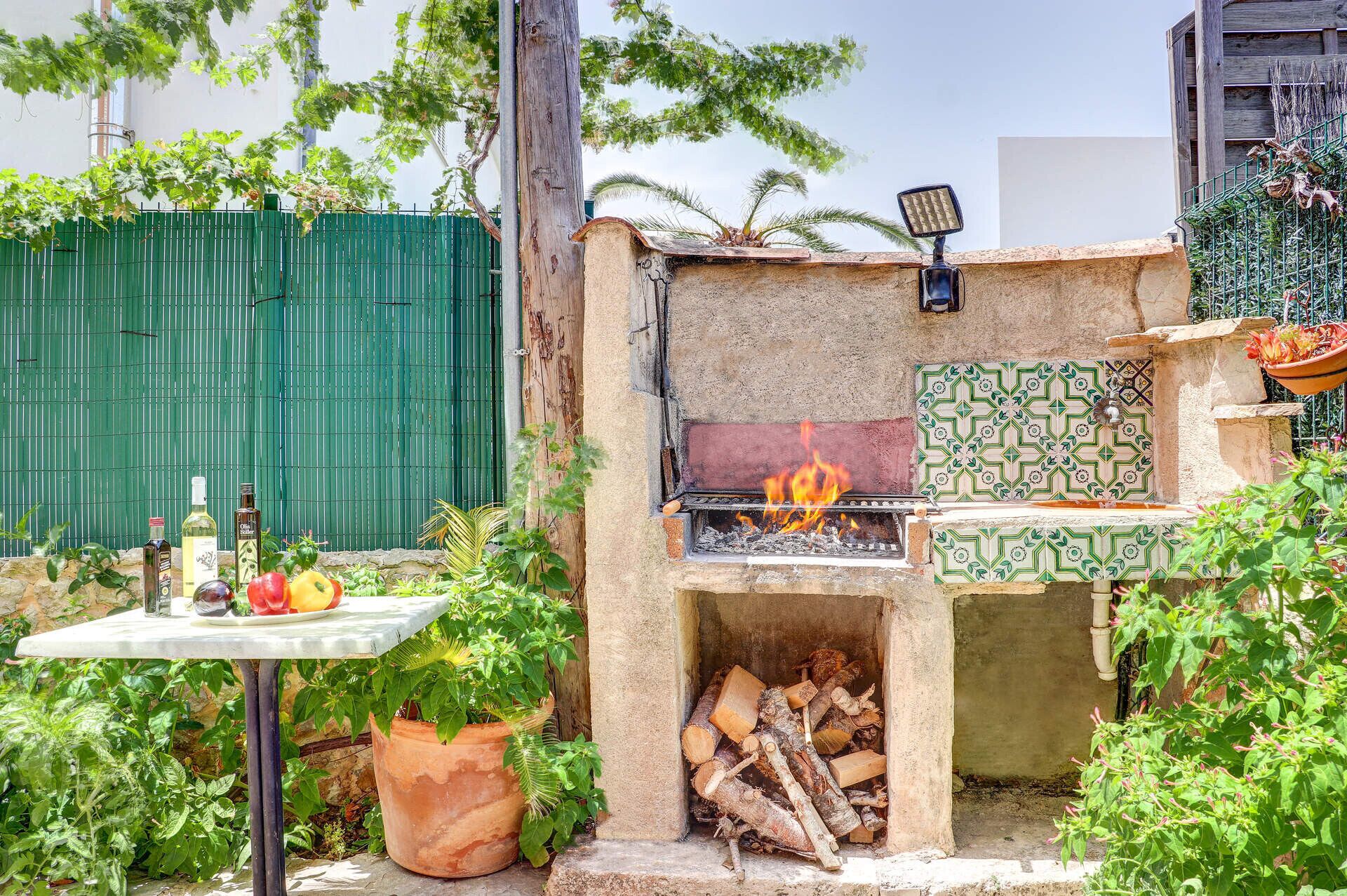 Un espace barbecue extérieur avec un four en pierre, une table avec des bouteilles et une clôture verte en arrière-plan.
