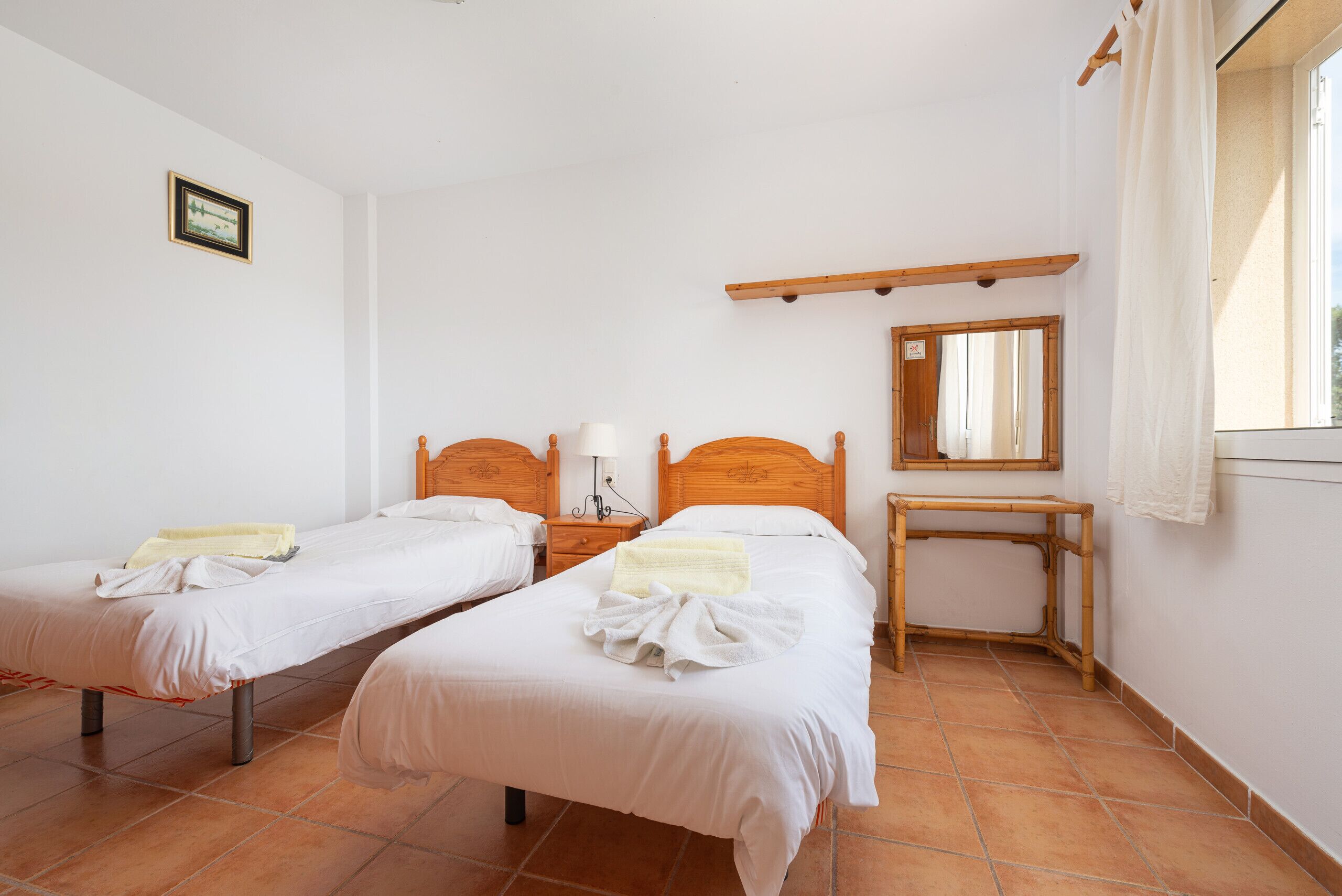Une chambre avec deux lits simples, une table de chevet en bois, une commode, un miroir et une fenêtre avec un rideau.