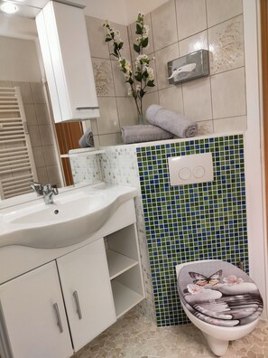Une salle de bain avec un lavabo blanc, un miroir, des toilettes dotées d’un abattant décoratif et un mur recouvert de carreaux verts et bleus.