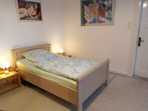 Quarto com cama de madeira, mesa de cabeceira com abajur e duas pinturas penduradas na parede.