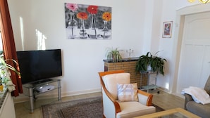 A living room with a flat-screen TV, a sofa, a chair, a coffee table, a patterned rug, and a painting of flowers on the wall. - Ferienhaus am See (Kirchlinteln)