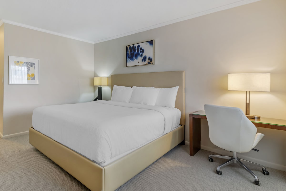 A hotel room with a bed, a desk, a chair, a lamp, and two wall art pieces.