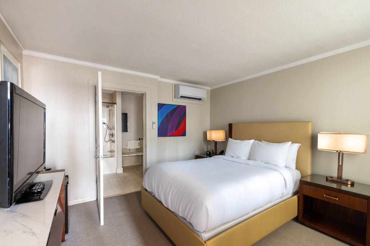 A hotel room with a bed, bedside tables, a TV, and an air conditioning unit.