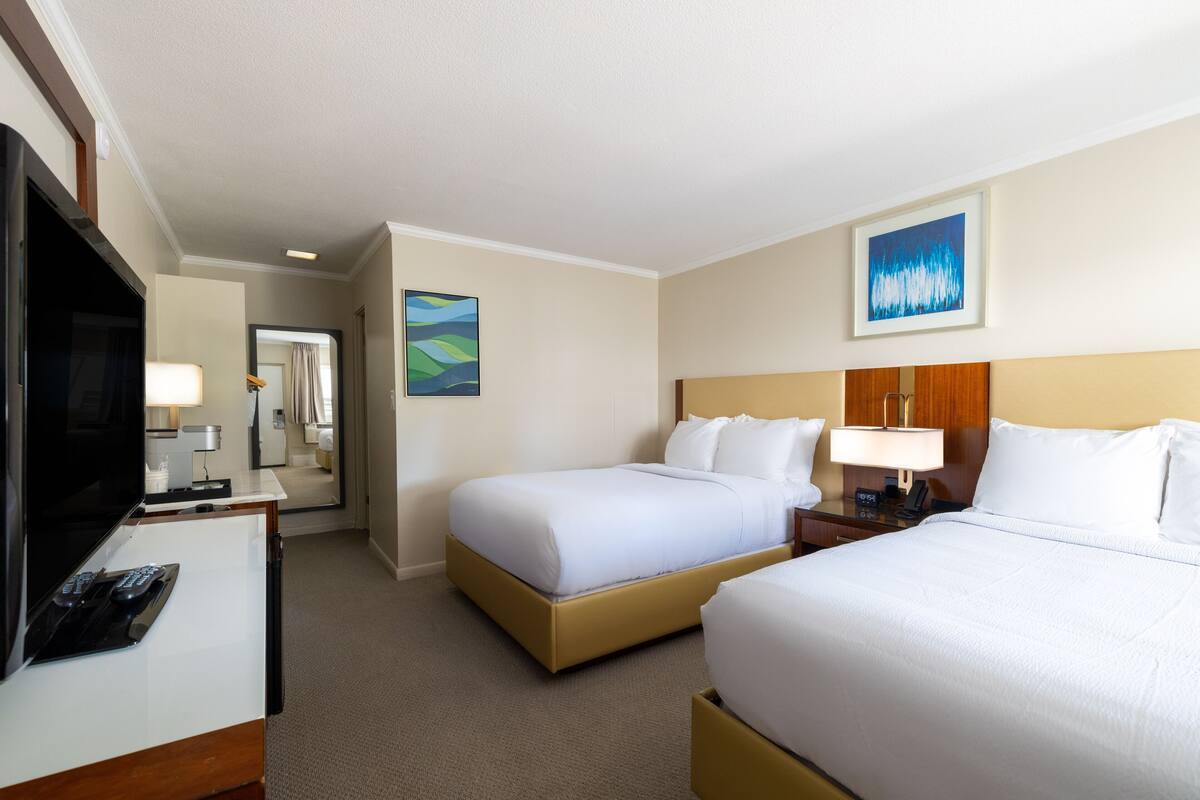 A hotel room with two beds, a desk with a TV and remote, a lamp, and a painting on the wall.