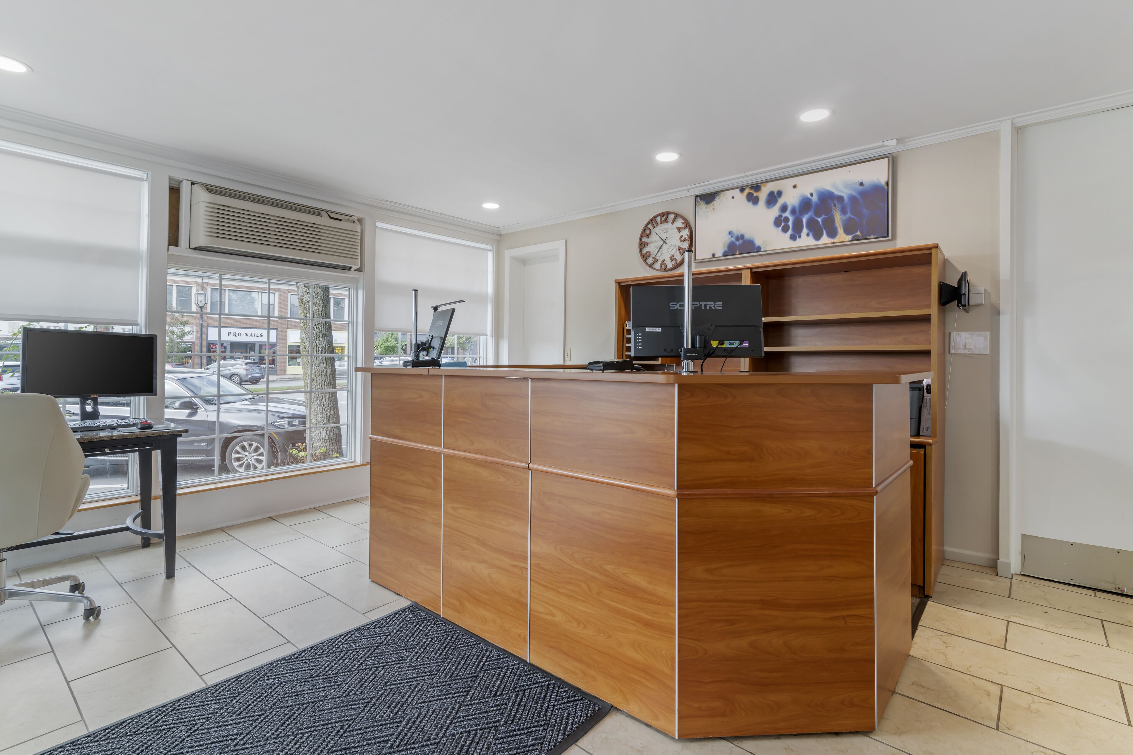 A reception desk with a computer, a clock, and a window view of a parking lot.
