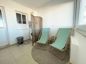 A small, well-lit room with a washing machine, a grey trash bin, and a patterned rug. There are two wooden chairs with green cushions. - Apartamento Acolhedor bem Localizado com Pátio Exterior no Centro de Lagos (Lagos)
