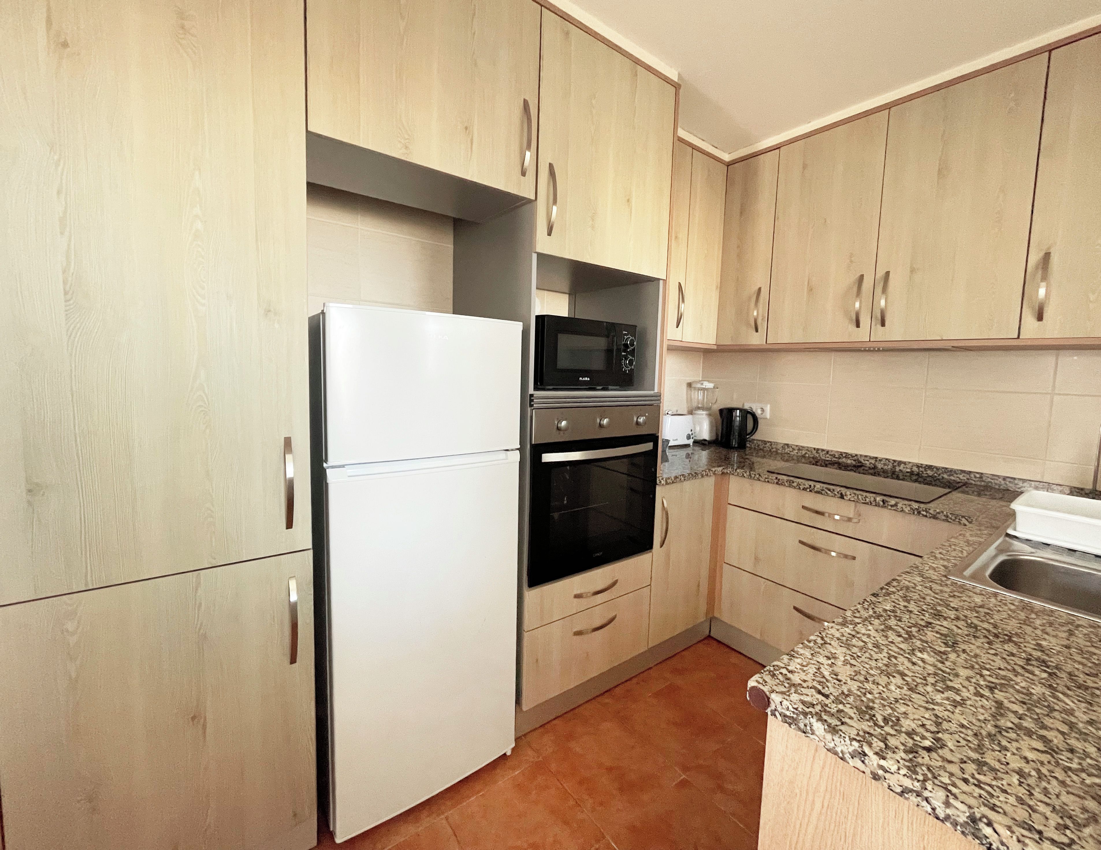 A kitchen with light wooden cabinets, a white refrigerator, a microwave, an oven, and a granite countertop.