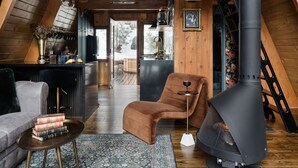 A cozy loft-style living space with a high ceiling, wooden beams, a fireplace, a sofa, a coffee table, and a rug. - Romantic A-Frame Cabin w/ Hot Tub, Fire Pit, Boulders, Fireplace, Hiking, Skiing (Running Springs)