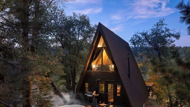 A cozy, triangular wooden cabin with a hot tub, deck, and outdoor seating area surrounded by trees. - Romantic A-Frame Cabin w/ Hot Tub, Fire Pit, Boulders, Fireplace, Hiking, Skiing (Running Springs)