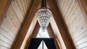 A cozy bedroom with a wooden ceiling, a large bed with a fluffy white blanket, a chandelier, and a view of trees through a window. - Romantic A-Frame Cabin w/ Hot Tub, Fire Pit, Boulders, Fireplace, Hiking, Skiing (Running Springs)