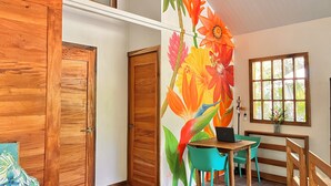 Une pièce avec des portes en bois, une fresque murale aux motifs floraux éclatants, une table en bois sur laquelle se trouve un ordinateur portable, des chaises de couleur turquoise et une fenêtre donnant sur un paysage verdoyant.