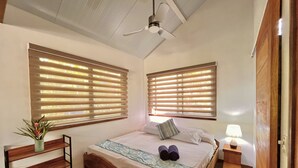 Une chambre avec un lit en bois, deux fenêtres équipées de stores, un ventilateur de plafond, une table de chevet avec une lampe et une petite plante posée sur une étagère.