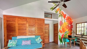 Une pièce avec un canapé en bois, une petite table, un ventilateur de plafond et une fresque murale aux motifs floraux éclatants.