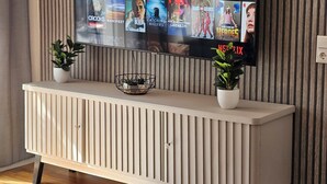 A flat-screen TV mounted on a wall with a Netflix interface displaying various shows. Below the TV, there is a white console with two potted plants. - The World Appartment (Magdeburg)