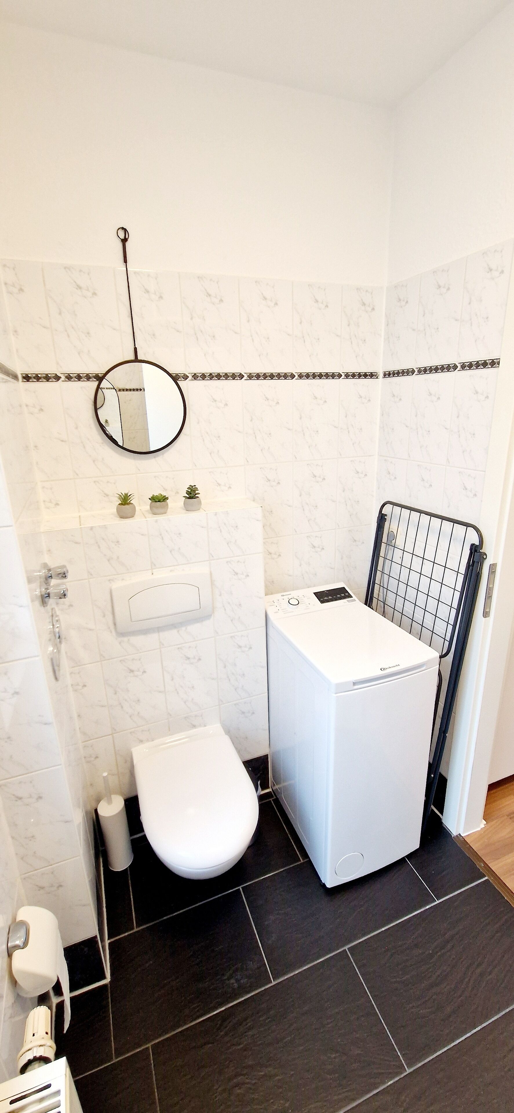 Une salle de bain avec des toilettes blanches, un lave-linge, un miroir et un porte-serviettes.