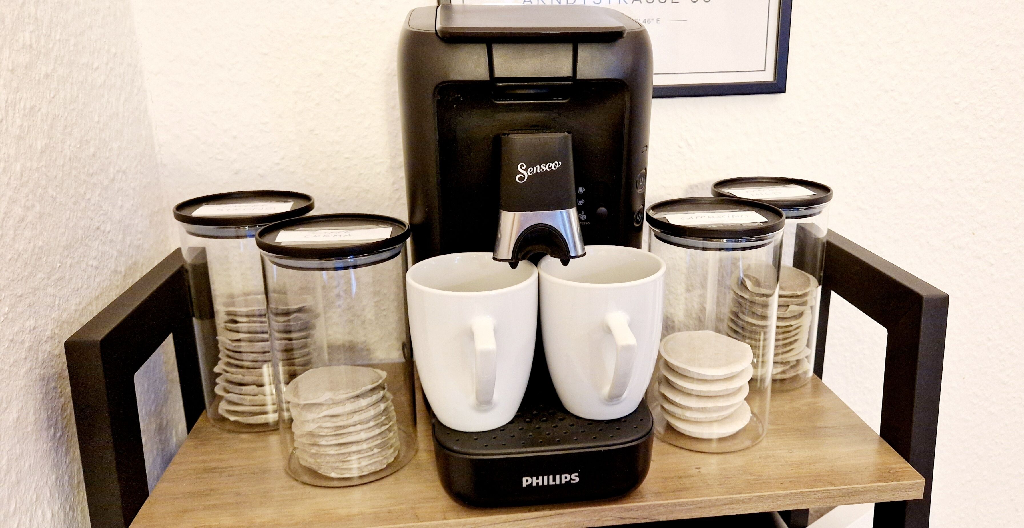 A coffee machine with two white cups, stacked plates, and clear jars on a wooden table.
