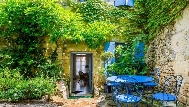 Un patio con piso de piedra, una mesa y sillas de metal azul, y una puerta que da a un espacio interior.