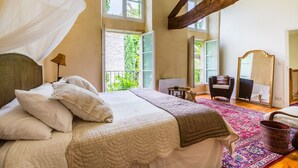 Un dormitorio con una cama grande, dosel, viga de techo de madera, una silla, una mesa, un espejo y vistas a zonas verdes.