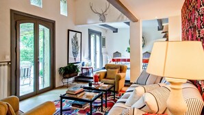 Amplia sala de estar con un sofá grande, una mesa de centro con libros, una lámpara y una chimenea. Se ve un comedor con mesa y sillas al fondo.