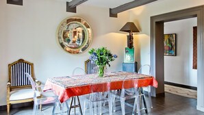 Un área de comedor con una mesa redonda cubierta por un mantel rojo, rodeada de sillas transparentes. Una silla de madera con un cojín a rayas está ubicada a la izquierda. Un espejo grande cuelga en la pared sobre la mesa, y un florero con flores está colocado sobre la mesa.
