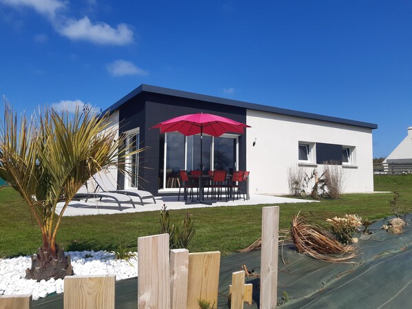 A modern house with a patio, a red umbrella, and a garden with plants and a palm tree.