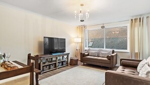 A living room with a flat-screen TV, a sofa, a coffee table, and a dining area with chairs and a wooden table.