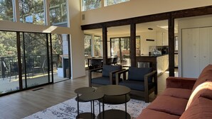 Un amplio salón con ventanas grandes, un rincón de estar acogedor y una cocina visible a través de un umbral abierto.
