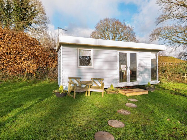 Une petite cabane d’un seul étage, à l’extérieur blanc, dotée d’une porte-fenêtre coulissante et d’une terrasse avec deux chaises en bois et une petite table.