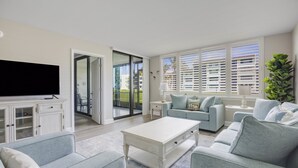 Ein geräumiges Wohnzimmer mit einem großen Flachbildfernseher, hellblauen Sofas, einem Couchtisch und einer Glasschiebetür, die auf einen Balkon führt.