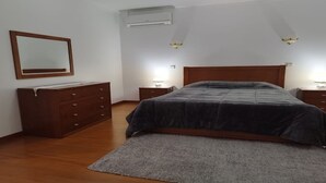 A bedroom with a wooden dresser, a bed with a dark blanket, two bedside lamps, and an air conditioning unit on the wall. - Casa Rural Muito Tranquila (Ortigosa de Cima)